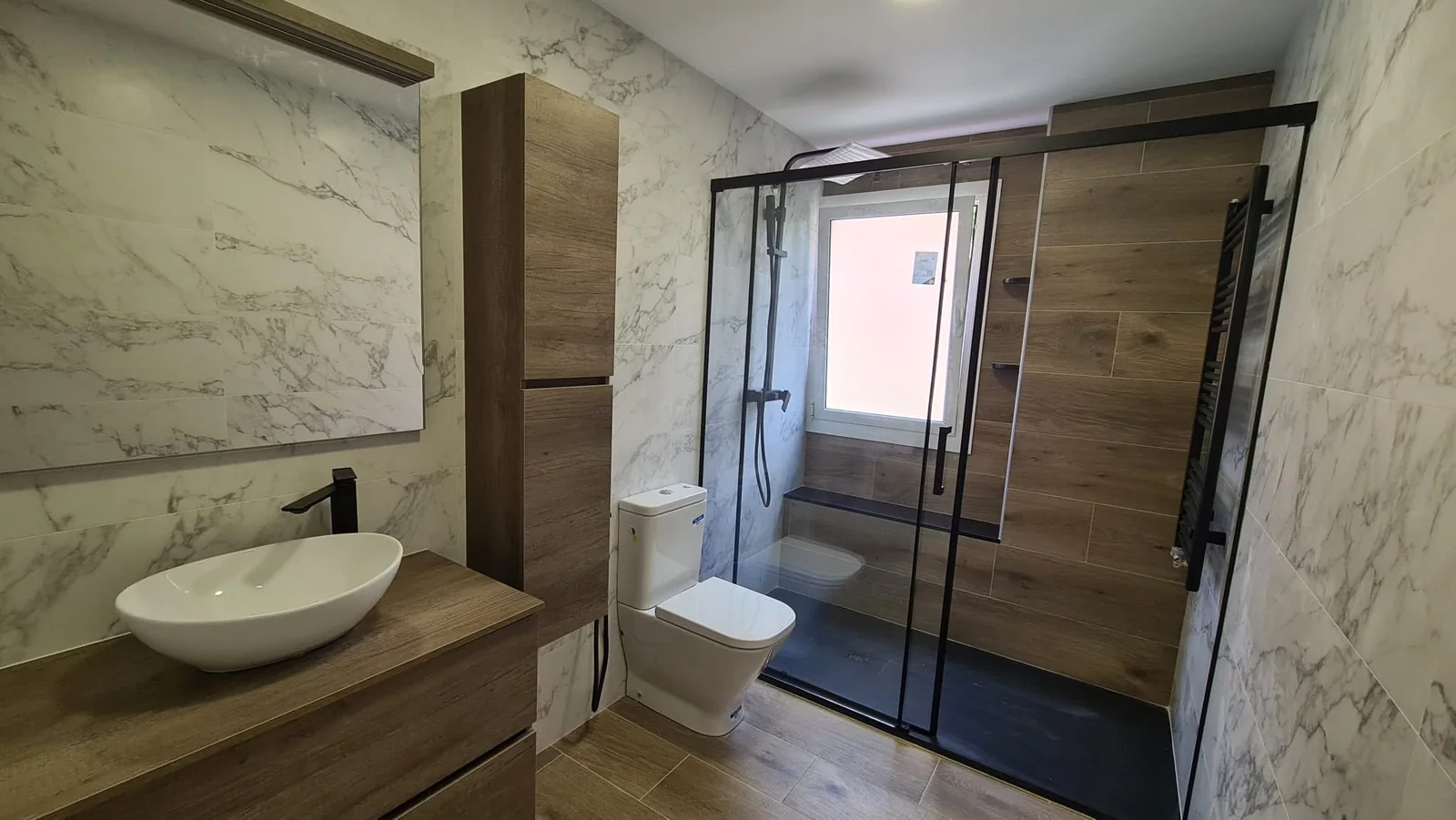Baño con mueble de madera, revestimientos claros y mampara de perfilería negra